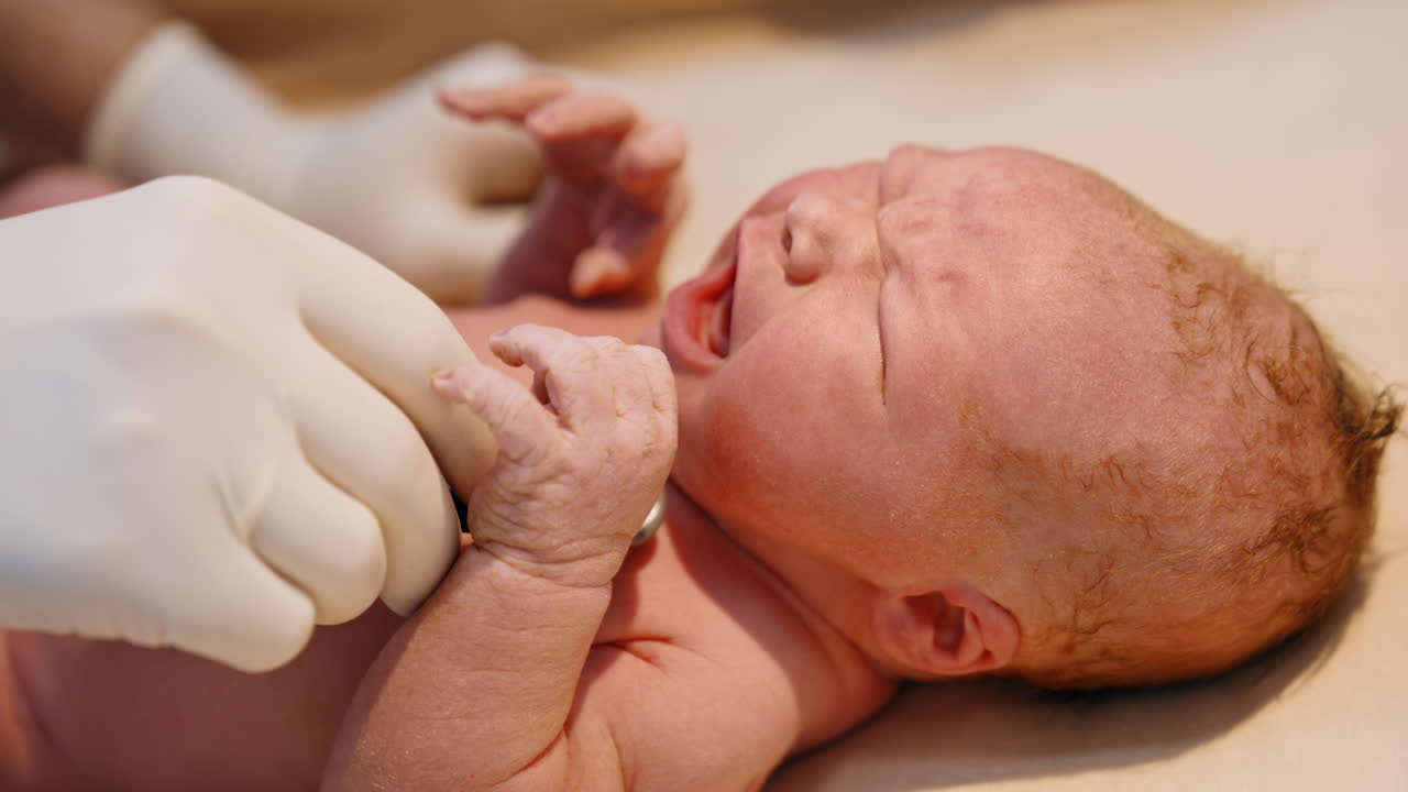 Newborn red-haired baby cries while doctor's gloved hand puts stethoscope to its chest. Checking the life of a child after birth. Close up.