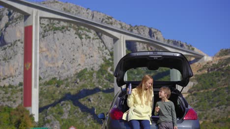 a man and his son are sitting and eating in their car with the moracica bridge in the background. they are taking a car trip through montenegro