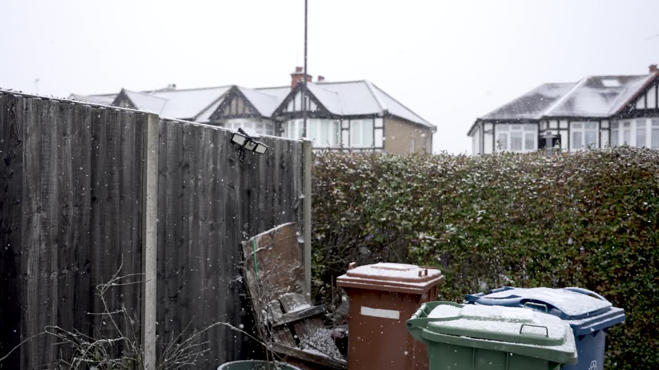 Neve che cade sui bidoni in un giardino anteriore a Londra, con una siepe e una recinzione visibili. La scena invernale mostra tetti innevati e un cielo nuvoloso in una zona residenziale.