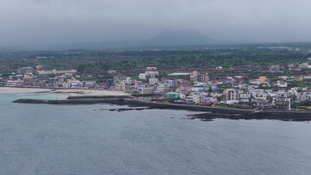 Inquadratura orizzontale a cameo con panoramica a sinistra mentre si avanza lungo la spiaggia di Gimnyeong sull'isola di Jeju, Corea del Sud, che mostra il villaggio costiero, l'oceano, le rocce vulcaniche e i cieli nuvolosi