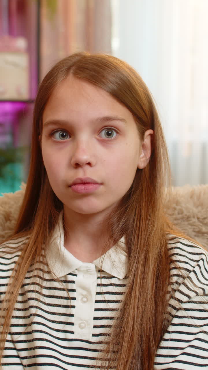 Child girl kid sitting on sofa at home showing silence gesture with finger near lips looking serious