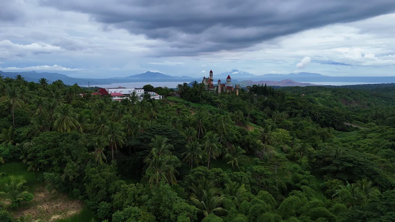 Aerial shot pushing past palm trees and moving towards a vibrant fantasy world in the Philippines, revealing whimsical architecture and lush surroundings under a cloudy sky
