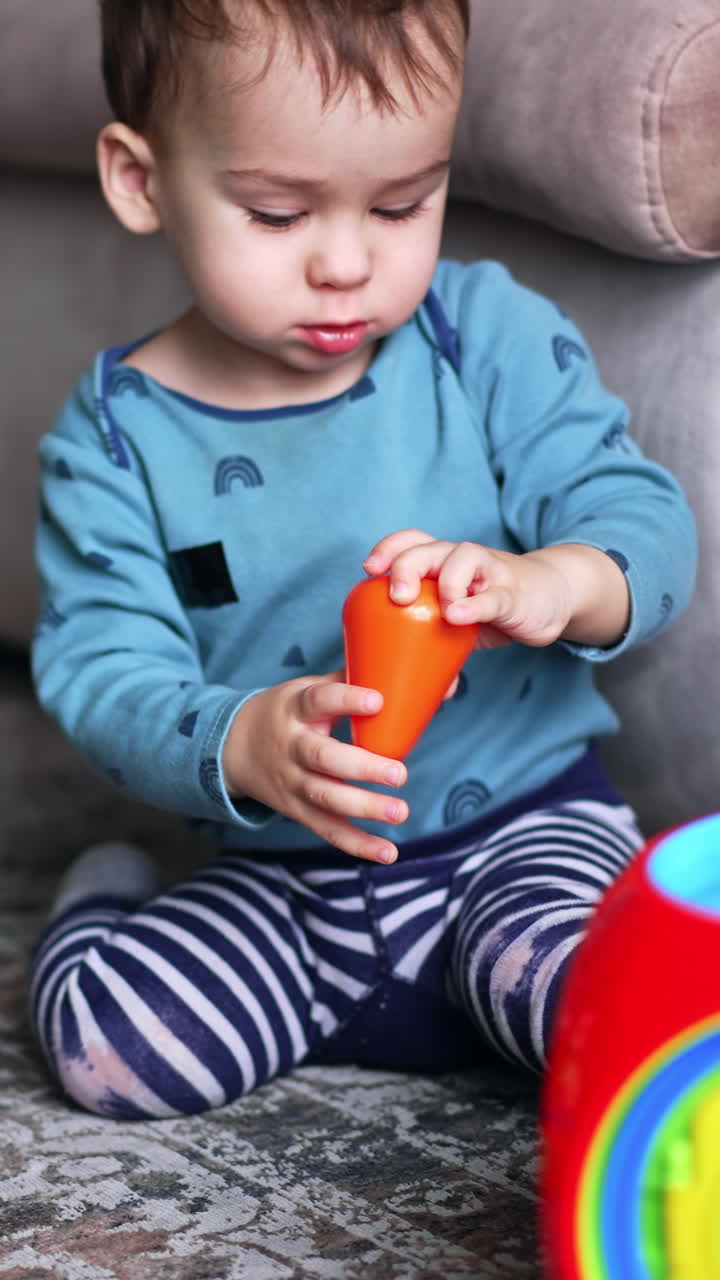 Dark-haired Caucasian toddler boy sitting on the floor next to the sofa. Adorable child plays with bright toys in front of him. Vertical video