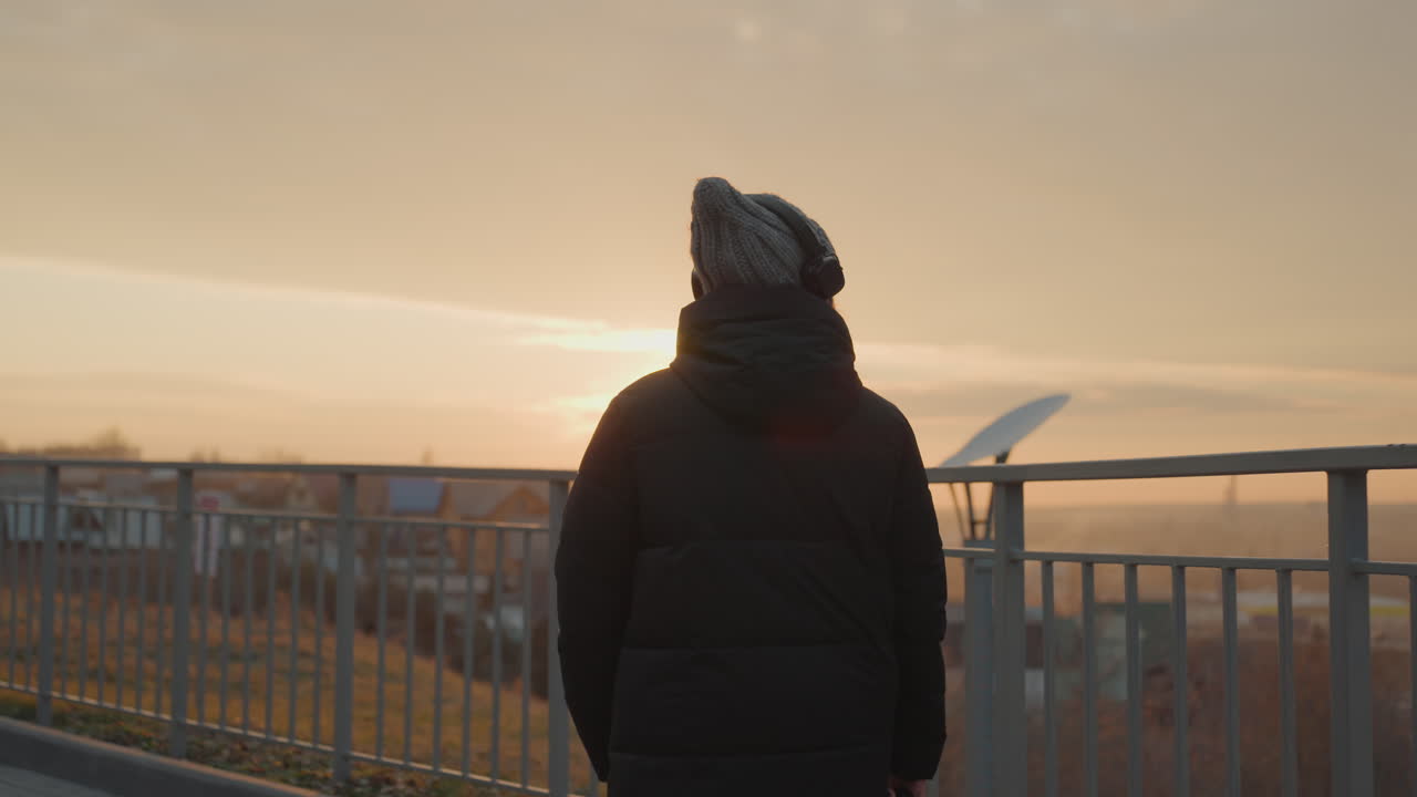 Kvinde i profil med høretelefoner ved solnedgang med udsigt over rækværk og by, langsom spadseretur langs promenade, varm gylden glød, eftertænksom stemning og blid bevægelse, mindfulness-vibe og filmisk indramning.