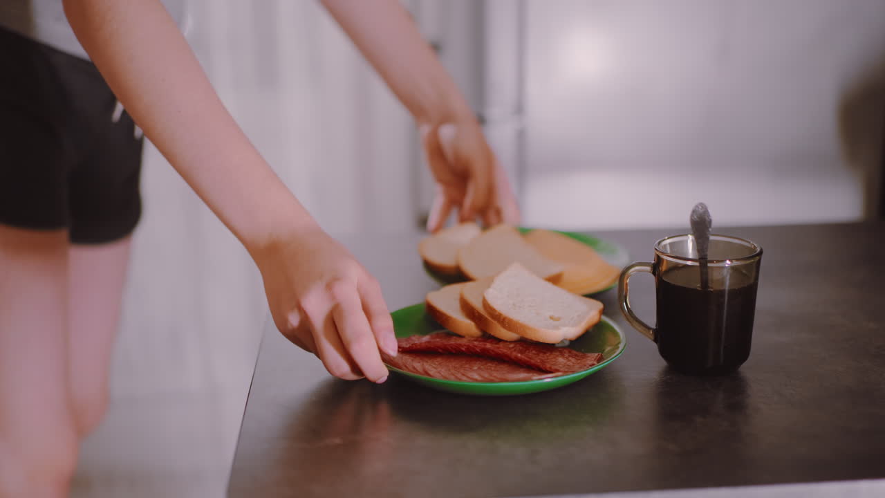 Lady places bread with meat on table near cup of coffee, capturing intimate breakfast preparation moment in kitchen atmosphere with natural light and casual lifestyle detail