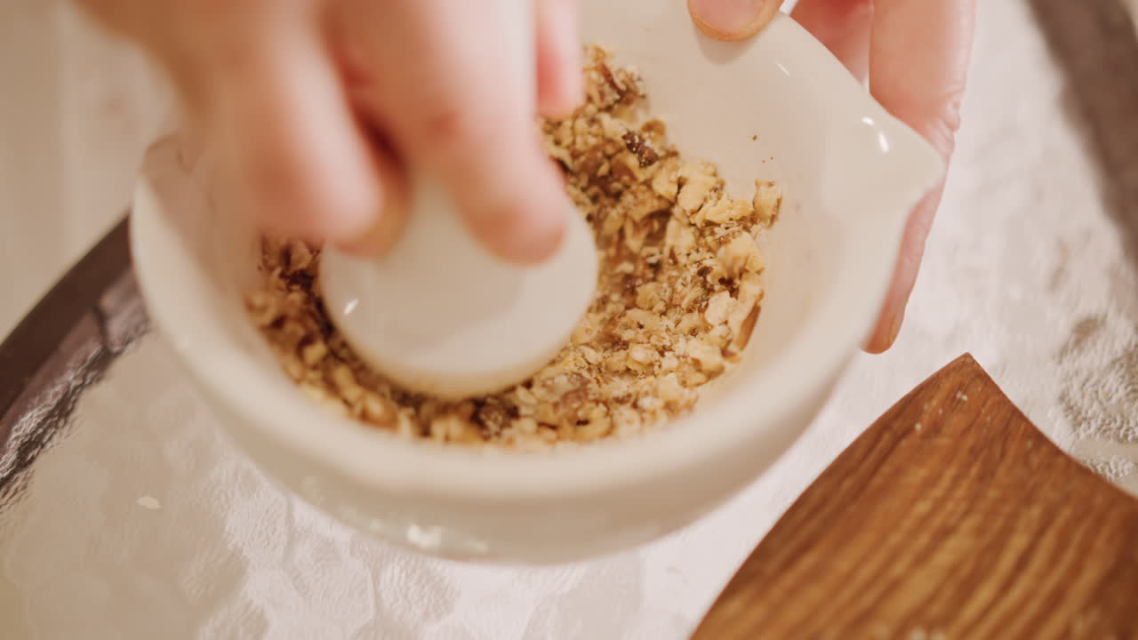 Primo piano di mani che tritano noci per il ripieno di una torta di zucca. Mortaio e pestello su un tavolo di pizzo, con il bordo di un tagliere di legno visibile. Una ciotola di ceramica rustica e una consistenza granulosa e friabile. La calda luce naturale evoca un'atmosfera di preparazione di dolci autunnale.