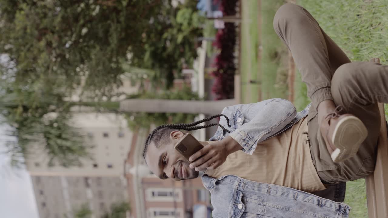 Man with braids relaxing on a park bench and using a smartphone