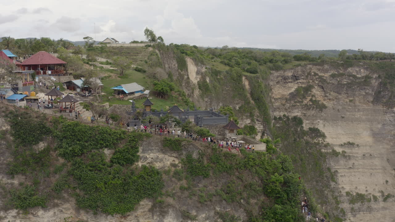 Kelingking beach scenic spot viewpoint with numerous tourists taking pictures on top of steep cliff on a cloudy day, Nusa Penida, Bali, Indonesia. Aerial shot