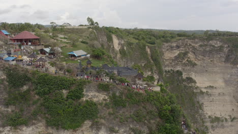 kelingking beach scenic spot viewpoint with numerous tourists taking pictures on top of steep cliff on a cloudy day, nusa penida, bali, indonesia. aerial shot