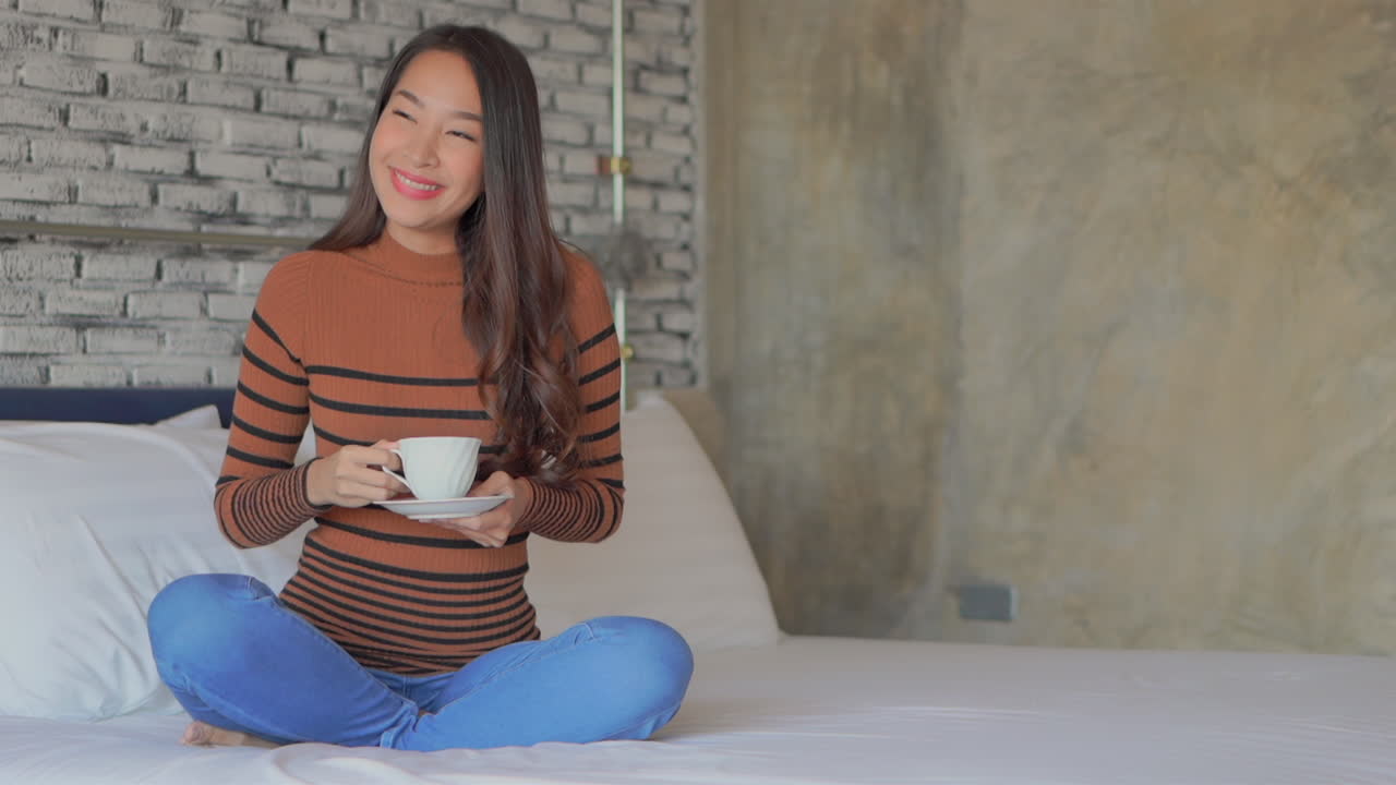 A pretty young Asian woman sitting on a bed sips a hot drink from a white coffee cup.copy space