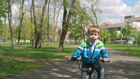 blond boy in sweatshirt and jeans riding a bike in the park 1