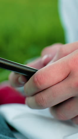 primer plano de la mano sosteniendo el teléfono inteligente y operándolo mientras está sentado en un campo cubierto de hierba, rodeado de exuberante vegetación, centrarse en los dedos y la pantalla del teléfono con una atmósfera al aire libre serena