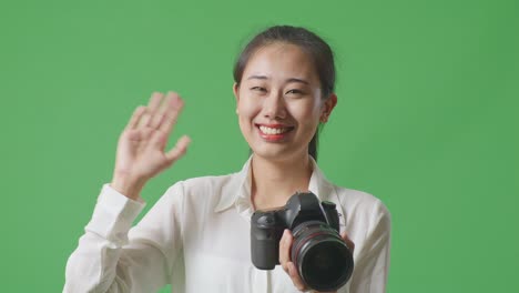 카메라에서 사진을 보는 아시아 사진작가의 클로즈업은 스튜디오에서 녹색 스크린 배경에 서서 손을 흔들고 있습니다.