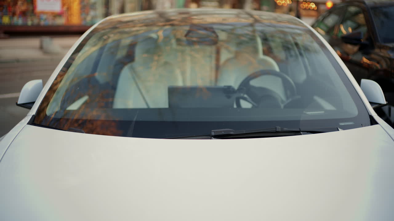 Vista frontale di un'auto elettrica bianca moderna parcheggiata su una strada cittadina con riflessi sul parabrezza.