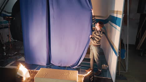 male technician pulls blue curtain during backstage television studio prep revealing news desk backdrop, floor lights blaze, cables trail, crane camera frames action, intense behind scenes production