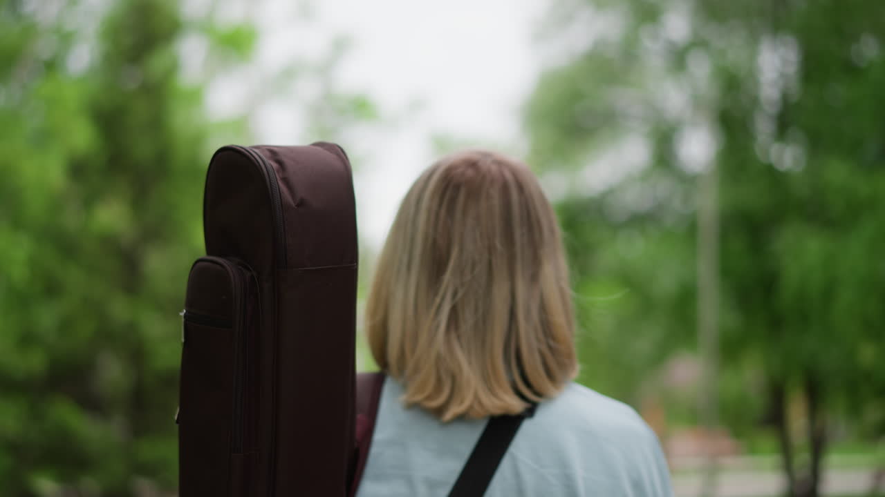 chitarrista caucasico con custodia per chitarra che cammina in un parco urbano alberato, giovane uomo biondo con custodia imbottita a tracolla, che guarda in alto e scruta l'orizzonte, ritratto spontaneo di musicista solitario