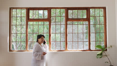 Mujer-Mayor-Afroamericana-Sonriendo-Mientras-Habla-Por-Teléfono-Inteligente-Cerca-De-La-Ventana-En-Casa