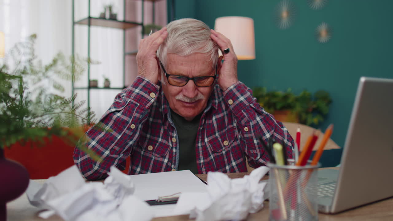 Senior man working on laptop computer, making notes on sheet of paper, upset by poor bad results