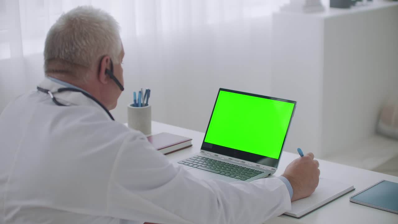 el médico masculino está consultando en línea desde su oficina en una computadora portátil con pantalla verde para la tecnología de llave croma mirando la pantalla
