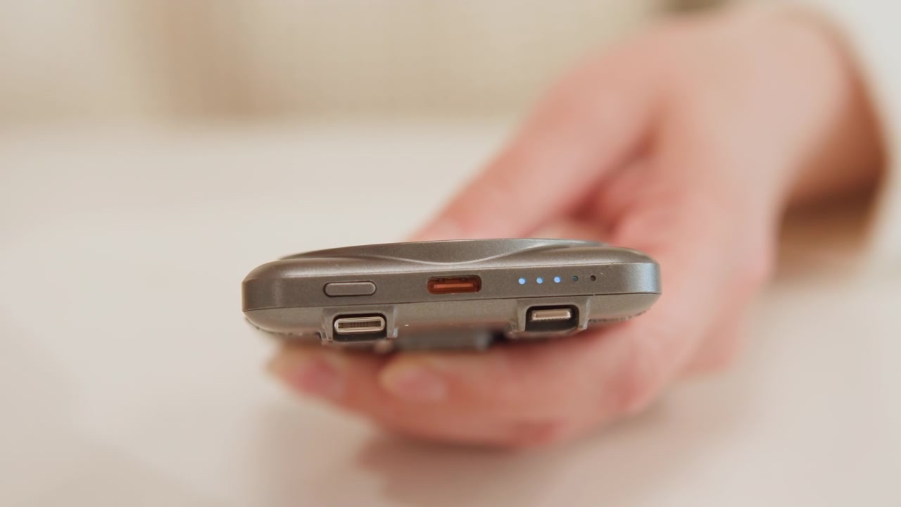 Nærbilde av en hånd som holder en liten håndholdt elektronisk fjernkontroll, med frontkanten med porter og knapper vendt mot betrakteren mens pekefingeren svever nær frontknappen under varmt innendørslys, noe som antyder bruker