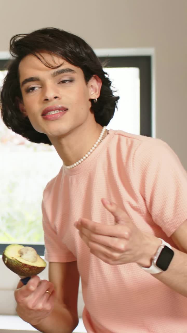 Vertical video: Facing camera, host teaching, holding halved avocado in kitchen, ring light moving