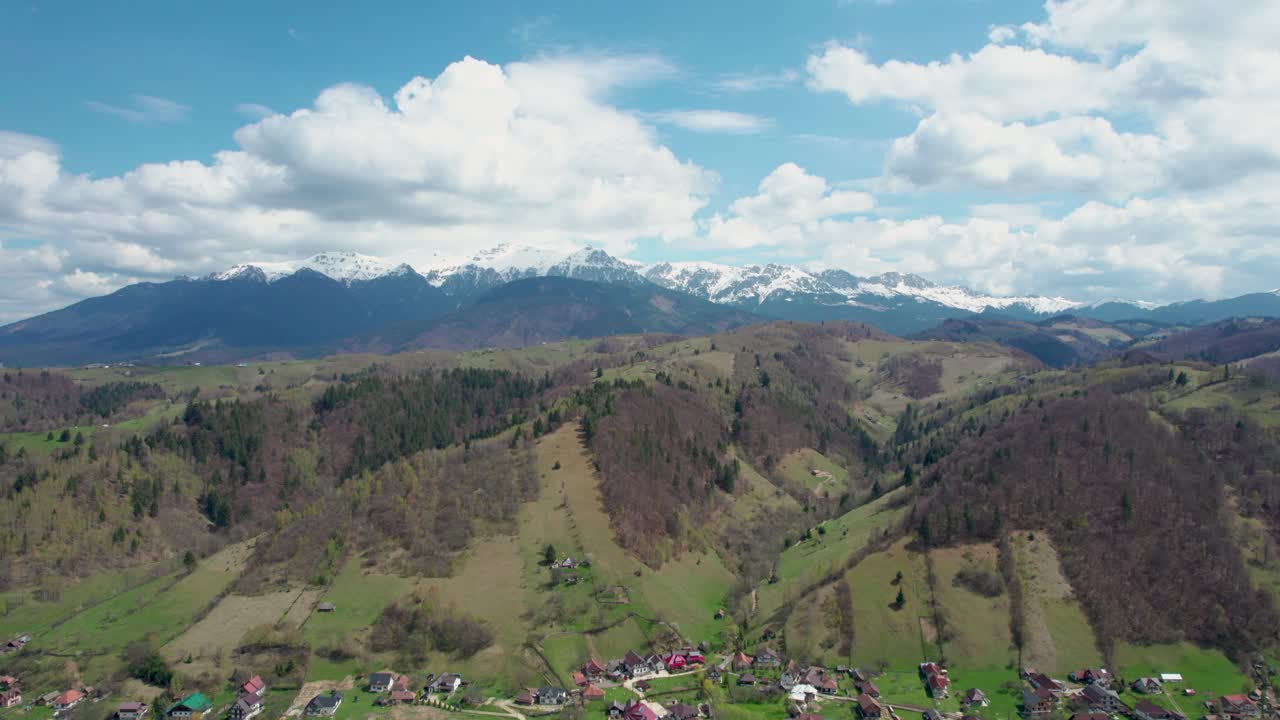 movimiento lateral del avión no tripulado con vistas a la cordillera de bucegi con picos nevados, cielo azul, gruesas nubes blancas y un pintoresco pueblo de montaña, rumania, transilvania, moieciu