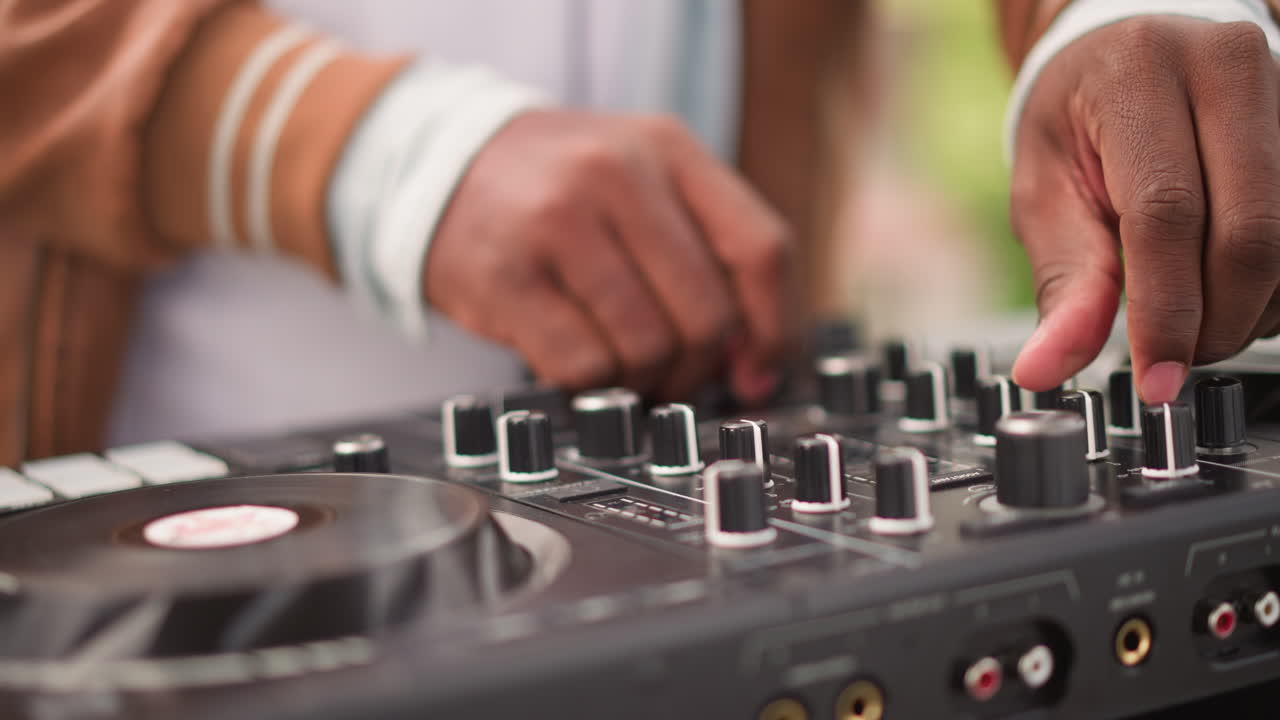 Primo piano della mano di un DJ nero che regola una manopola sul controller, atmosfera vivace di festa in giardino, luce del giorno nitida, cambi di tempo energici, jog wheel in vinile e movimenti tattili dei fader, animatore informale che si connette