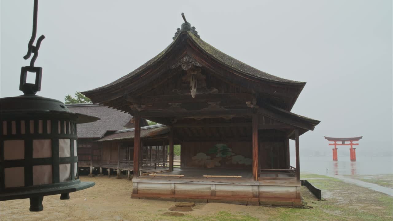Rainy day shot of Miyajima’s Itsukushima Shrine stage with a distant view of the iconic torii gate. A hanging lantern adds foreground depth.