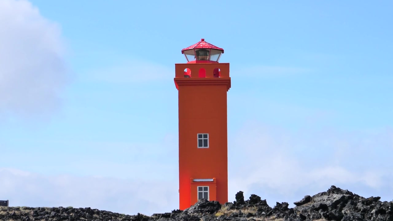 Udsigt fra jordhøjde af Svörtuloft Fyr på den vestlige kant af Snaefellsnes-halvøen i Island, med rødt fyr, vulkanske klipper, blå himmel og kystlandskab.