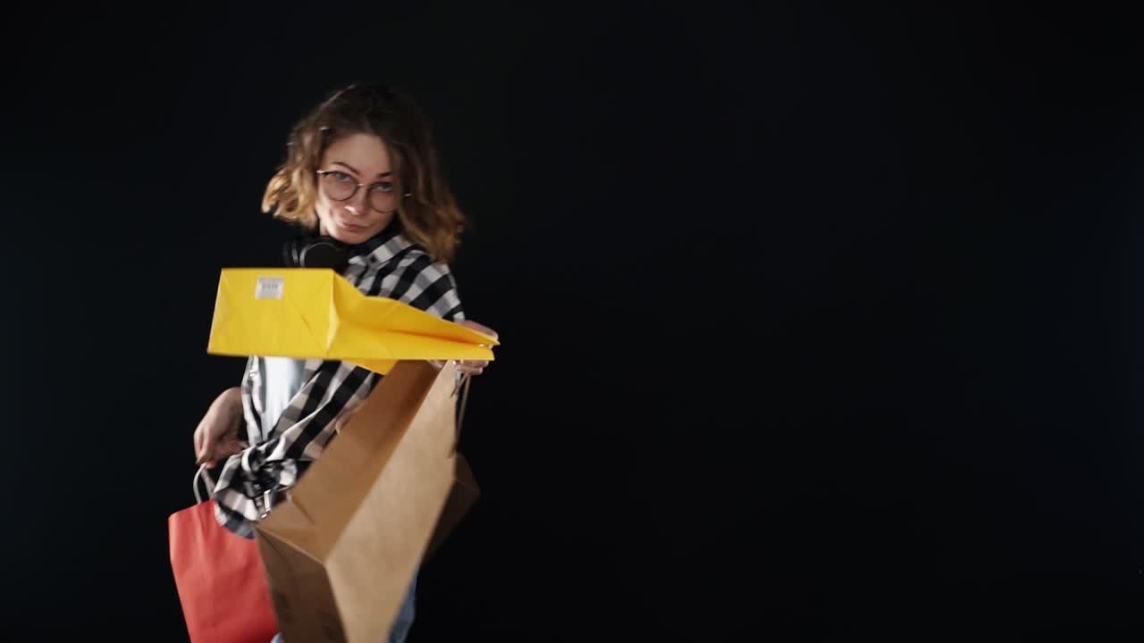 검은 배경 위에 다채로운 쇼핑 가방과 함께 행복하게 걸어 아름다운 미소 여성 plaid 셔츠와 청바지. 목에 안경과 검은 헤드폰을 착용