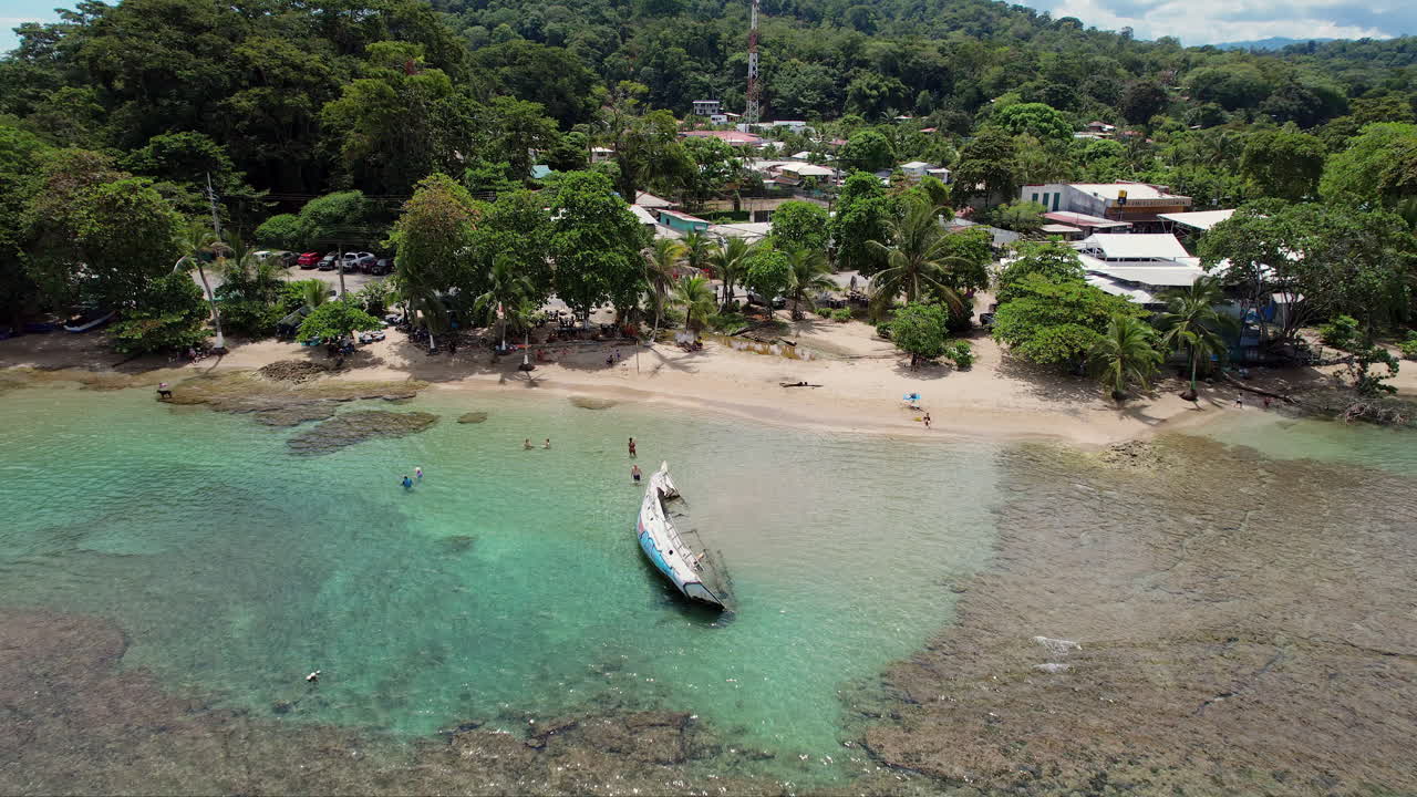 A panning overhead drone shot of a shipwreck in sparkling blue waters off the beautiful coast of Puerto Viejo, Costa Rica as snorkelers swim and people walk on the beach under palm trees nearby.