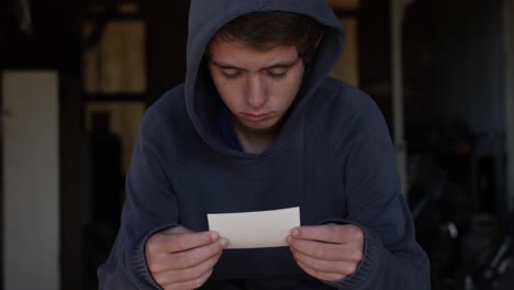 a sad and depressed looking teenage boy wearing a blue hooded shirt sits alone in a dirty garage looking at a picture