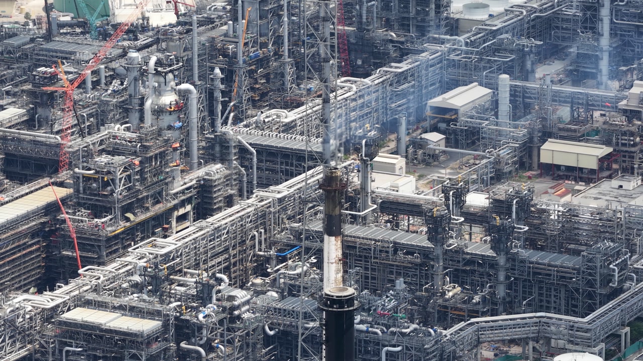 High aerial view of a huge oil refinery with complex steel pipelines and structures emitting steam, showcasing the scale of the energy and manufacturing industry for fuel and gas production