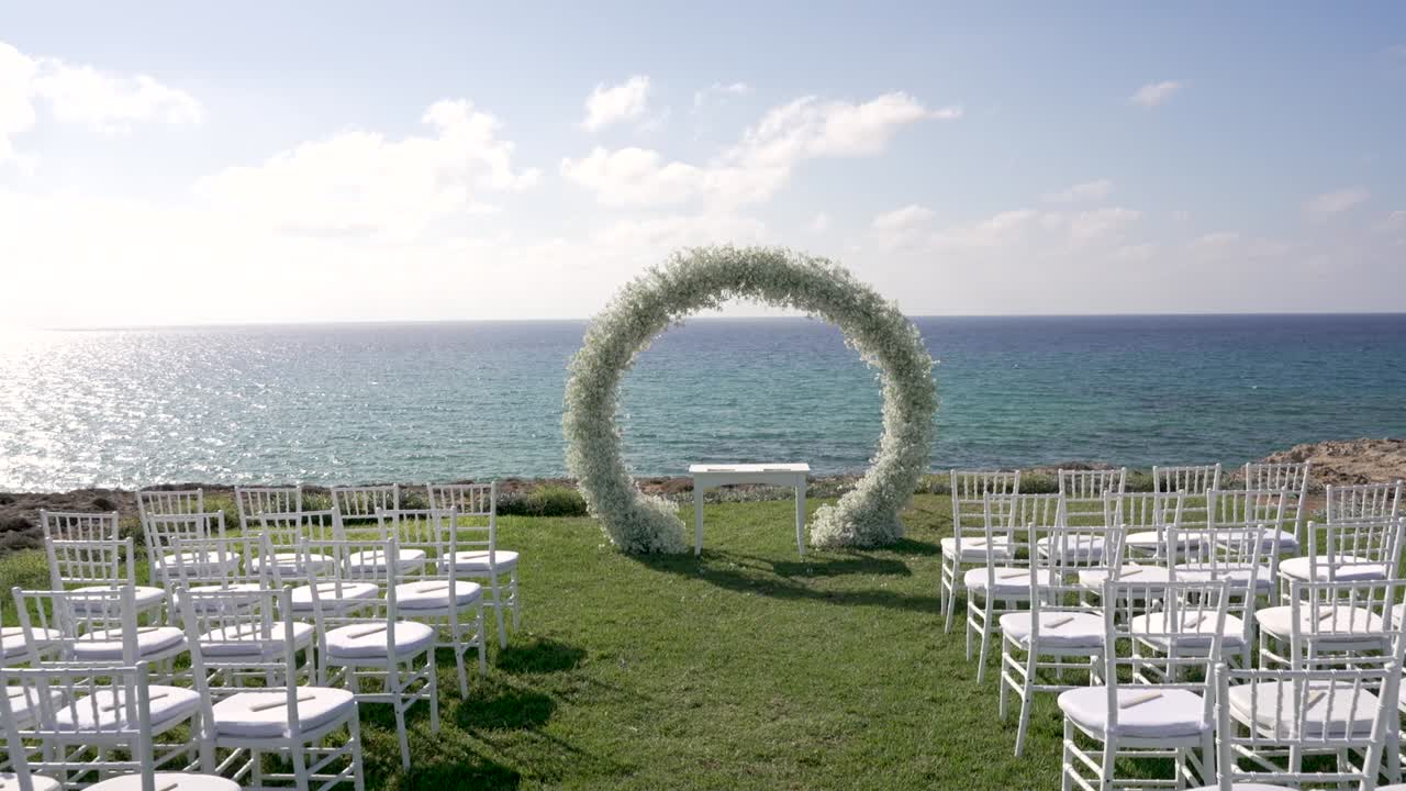 Lungo la costa è visibile l'allestimento di un matrimonio. Delle sedie vuote sono disposte di fronte a un arco floreale, con vista sull'oceano sotto un cielo sereno. La scena cattura i preparativi per la cerimonia.