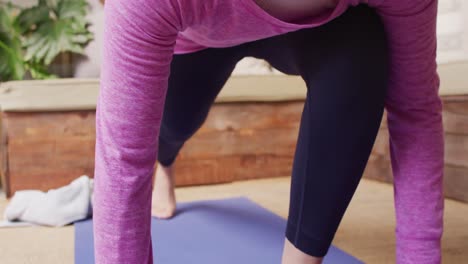 Video-of-relaxed-biracial-woman-in-hijab-stretching-leg-in-yoga-pose-kneeling-on-mat-in-living-room