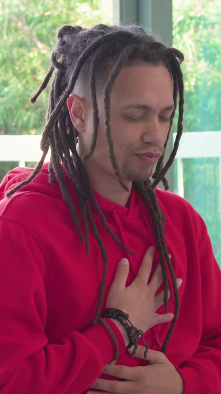 Vertical video: Closing eyes man in red hoodie placing hands on chest by balcony, grounding breath