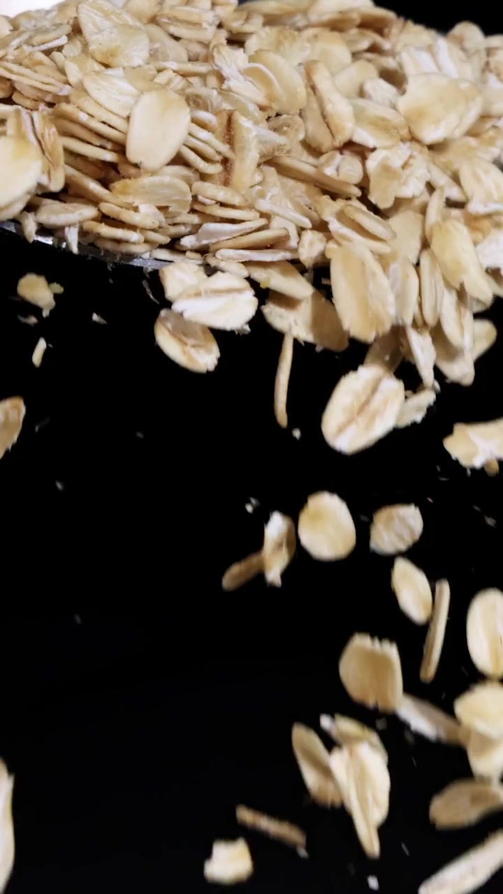 Fiocchi d'avena cadono da un cucchiaio al rallentatore, su uno sfondo scuro, evidenziandone la consistenza e il movimento.