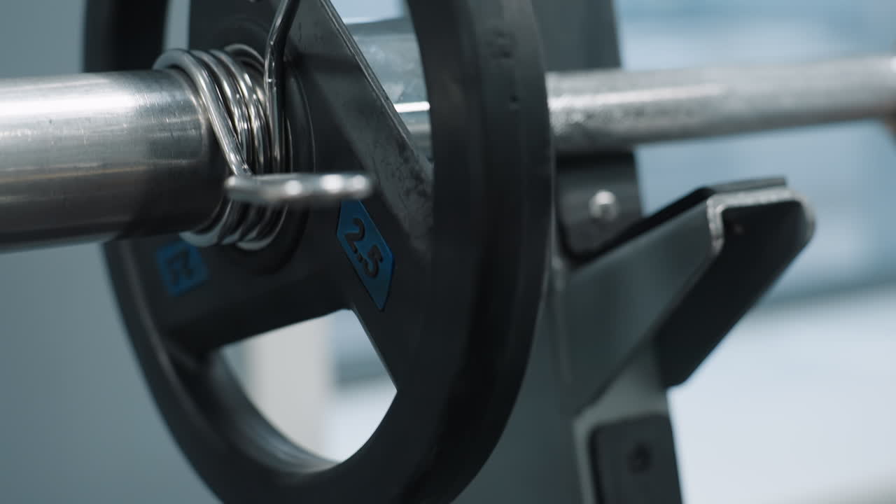 Close up of metal weight plate secured with clip as it drops onto iron rack in gym environment, barbell resting on holder, steel textures and gym setup visible in frame with shallow depth of field