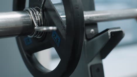 close up of metal weight plate secured with clip as it drops onto iron rack in gym environment, barbell resting on holder, steel textures and gym setup visible in frame with shallow depth of field