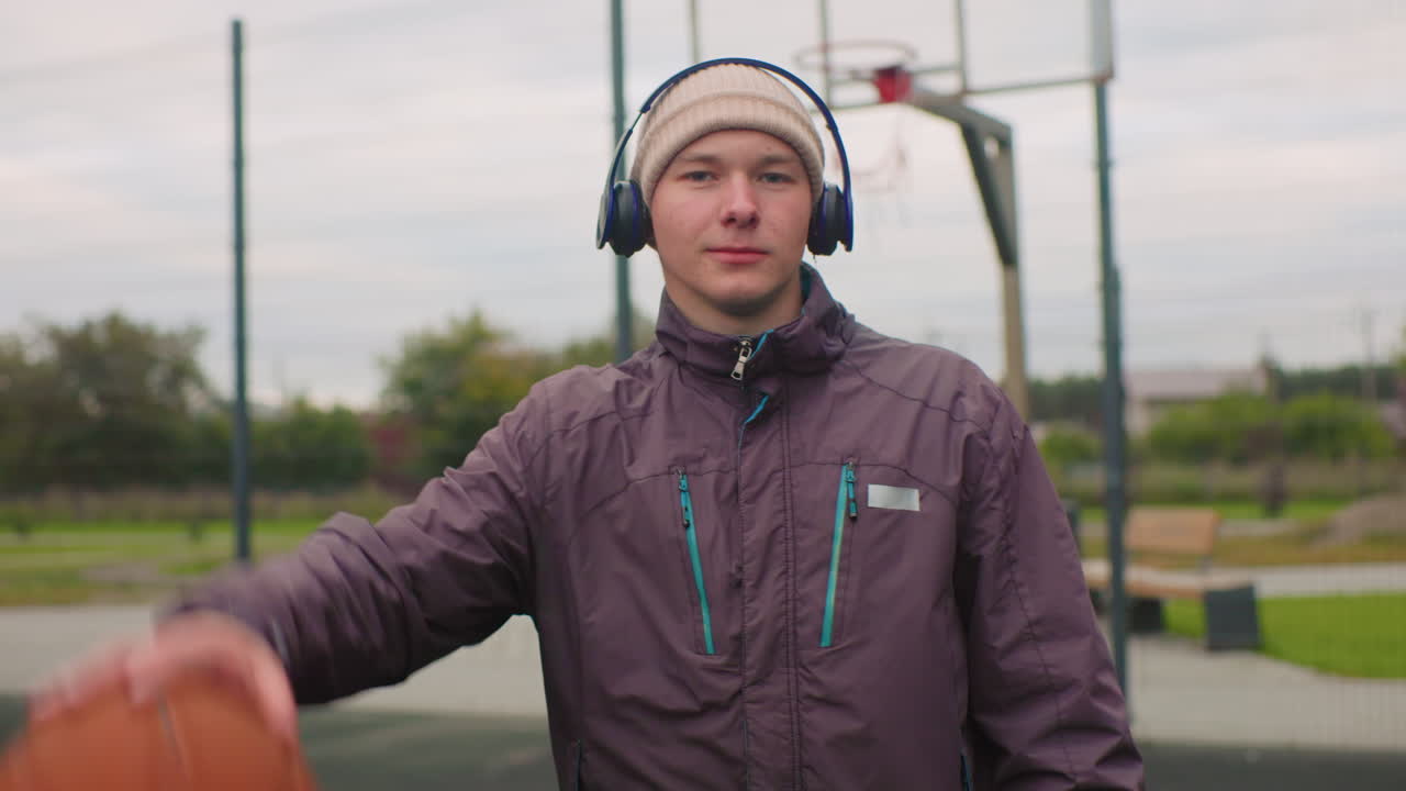 young man on basketball court wearing headphones, layered jacket and beanie, casually dribbling basketball, posing for portrait under overcast sky, hoop and park in background, relaxed urban sports