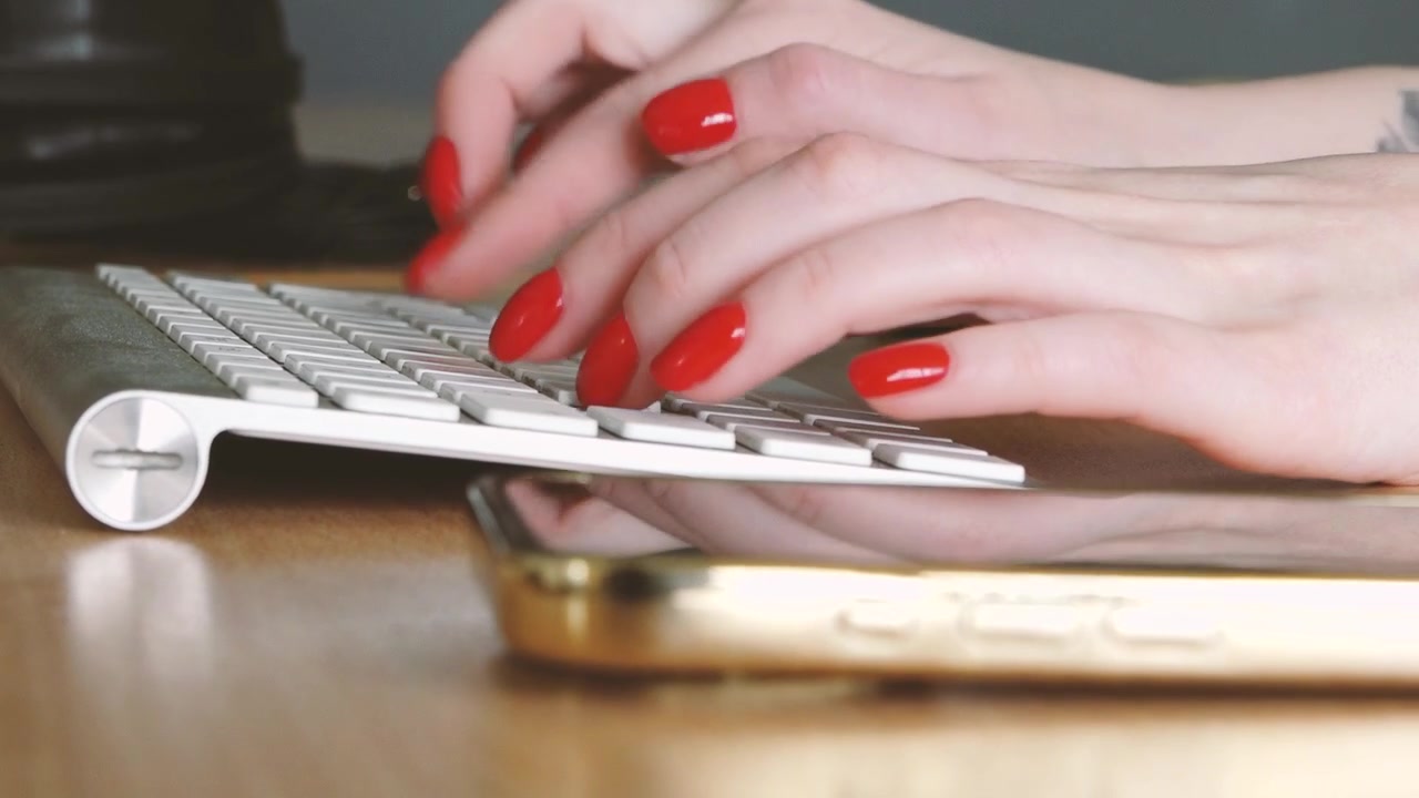 Close-up of hands with red nails typing on a slim keyboard, with a smartphone lying on a wooden desk in a modern workspace.