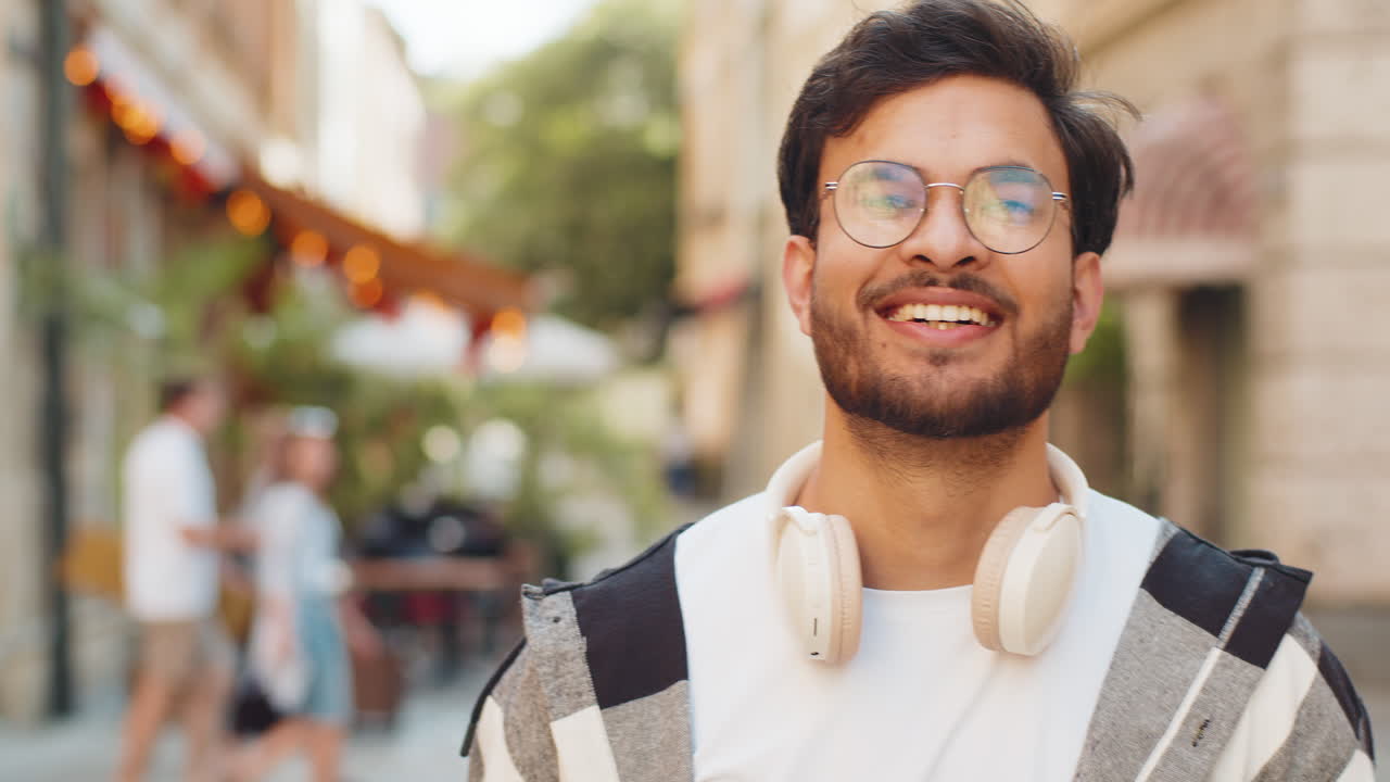 Happy arabian man taking off wireless headphones and looking at camera smiling in city street