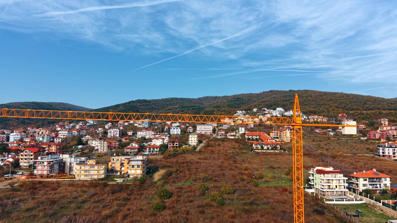 Vista aerea di una città costiera con un'alta gru da costruzione arancione in primo piano. Una gru a torre arancione si erge sopra una città costiera collinare mentre la macchia tardo autunnale copre il pendio sotto colline boscose.