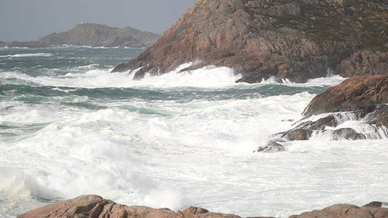 Waves violently crash against rugged rocks, sending white foam into the air. The coastal scene features a dramatic overcast sky, creating a moody atmosphere