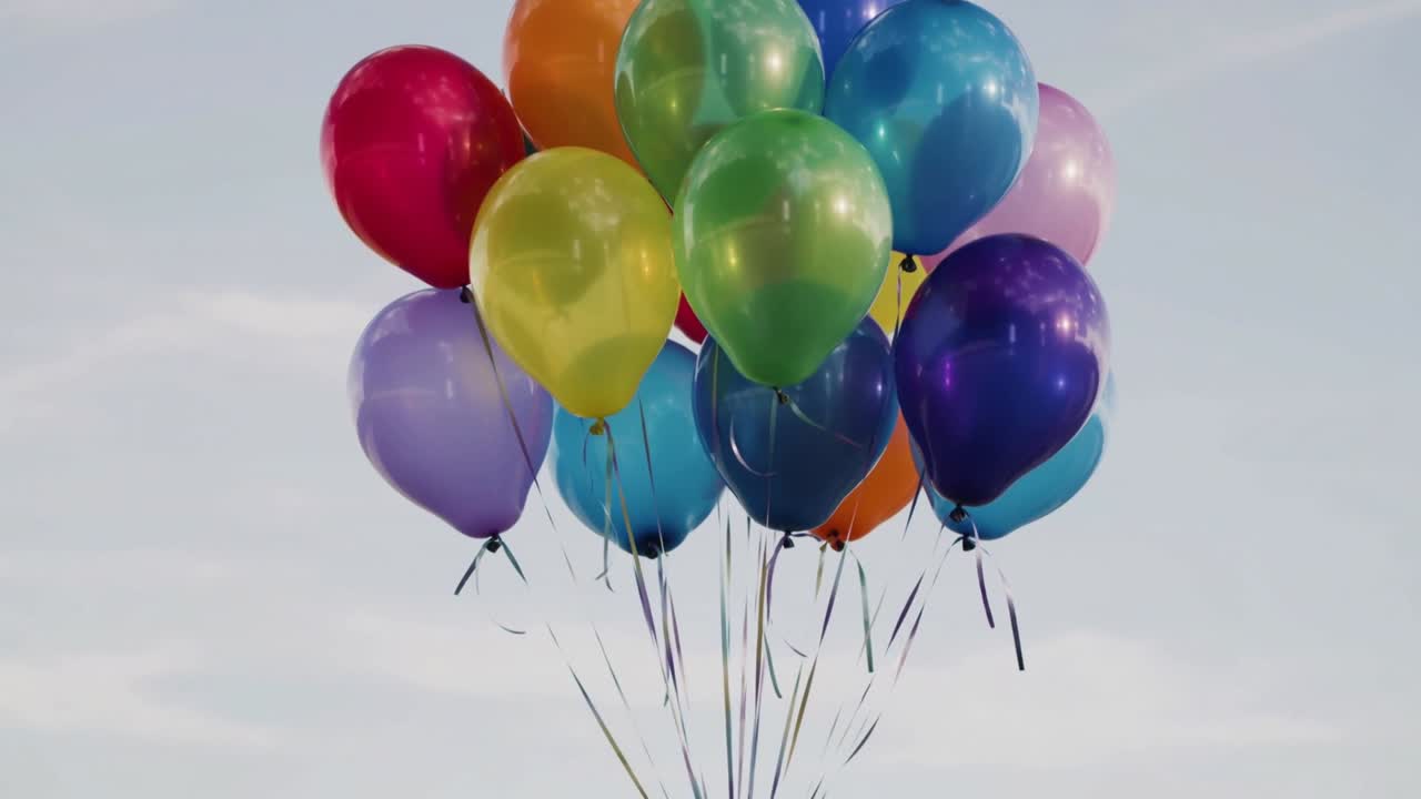 Pohupující se shluk 14 heliových balónků unášených modrou oblohou, stuhy vlající ve vánku.