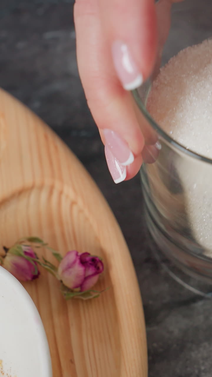 primer plano de una persona colocando suavemente un vaso transparente lleno de azúcar blanco en una mesa negra elegante, vista parcial de una bandeja de madera con especias y flores secas, junto con otros artículos esenciales de la cocina
