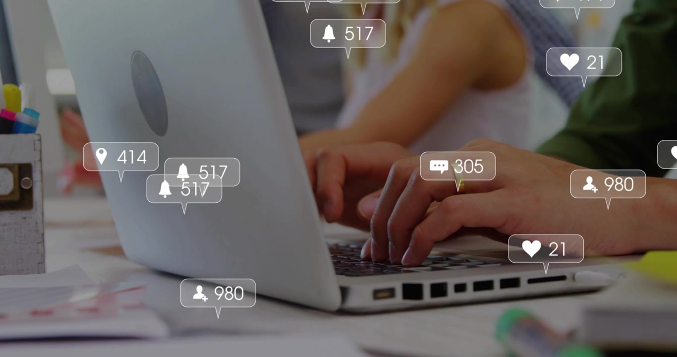 Creators typing on laptop in shared desk, hands pressing keys causing social media icons rising