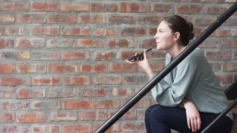 Young-Caucasian-businesswoman-records-a-voice-message-on-her-phone,-seated-on-stairs,-with-copy-spac