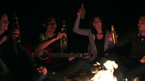 Group-of-young-people-near-campfire-drinking-beer-and-clicking-bottles-with-cheers-late-at-night.-Slow-Motion-shot