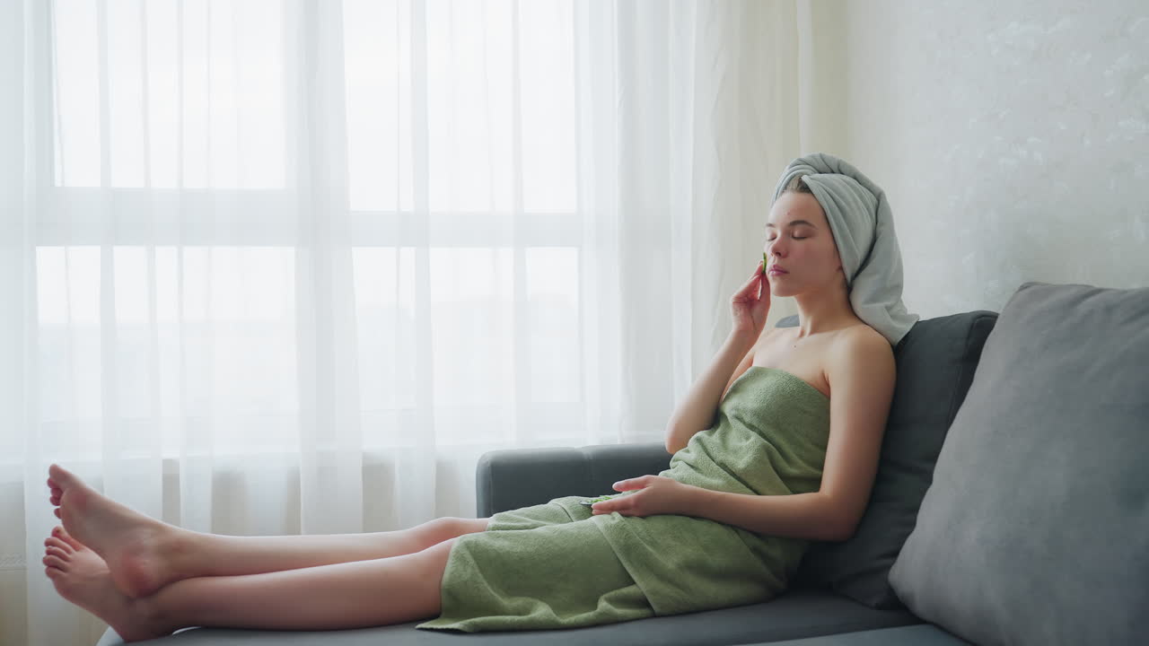 Woman sitting on couch with legs stretched out, wrapped in green towel and towel on head, gently placing cucumber slices on cheeks as part of skincare routine in peaceful naturally lit room